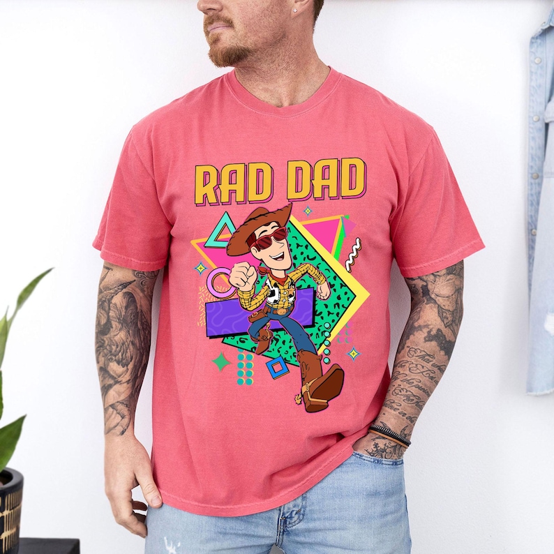 May include: A coral-colored t-shirt featuring the phrase "RAD DAD" in yellow and orange block letters. The shirt displays a cartoon image of a cowboy character with sunglasses, set against a geometric background with vibrant colors.