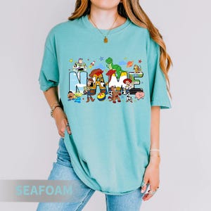 Puede incluir: Camiseta verde agua con la palabra "NAME" en un dise&ntilde;o colorido con personajes de dibujos animados. La camiseta tiene mangas cortas y un corte relajado. El dise&ntilde;o incluye personajes de una popular pel&iacute;cula de animaci&oacute;n.