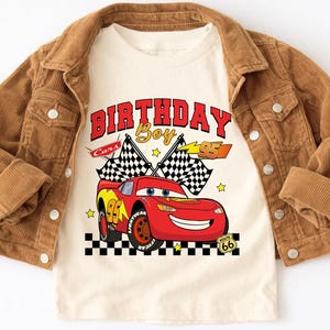 May include: A tan t-shirt featuring a red race car graphic with the text "BIRTHDAY Boy" and the number "95". The shirt is layered under a brown corduroy jacket.