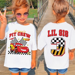 May include: White t-shirt with a graphic of a red race car and the text "Prince's PIT CREW" on the front. The back of the shirt has the text "LIL SIS" above a number 3 graphic. The shirt is worn by a young child.