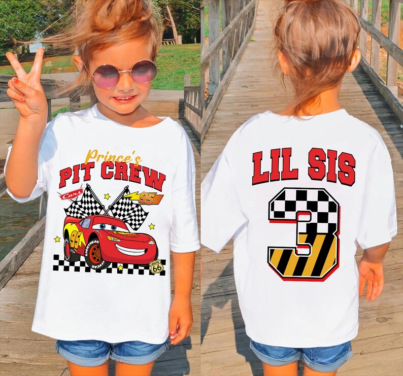 May include: White t-shirt with a graphic of a red race car and the text "Prince's PIT CREW" on the front. The back of the shirt has the text "LIL SIS" above a number 3 graphic. The shirt is worn by a young child.