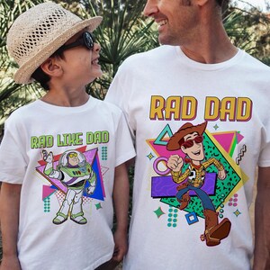 May include: White t-shirts with cartoon graphics. One shirt says "RAD DAD" with a Woody character, the other says "RAD LIKE DAD" with a Buzz Lightyear character. Both shirts have colorful geometric designs.
