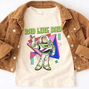 May include: A tan t-shirt with the text "RAD LIKE DAD" in green and a cartoon image of Buzz Lightyear. The shirt is layered under a brown corduroy jacket.