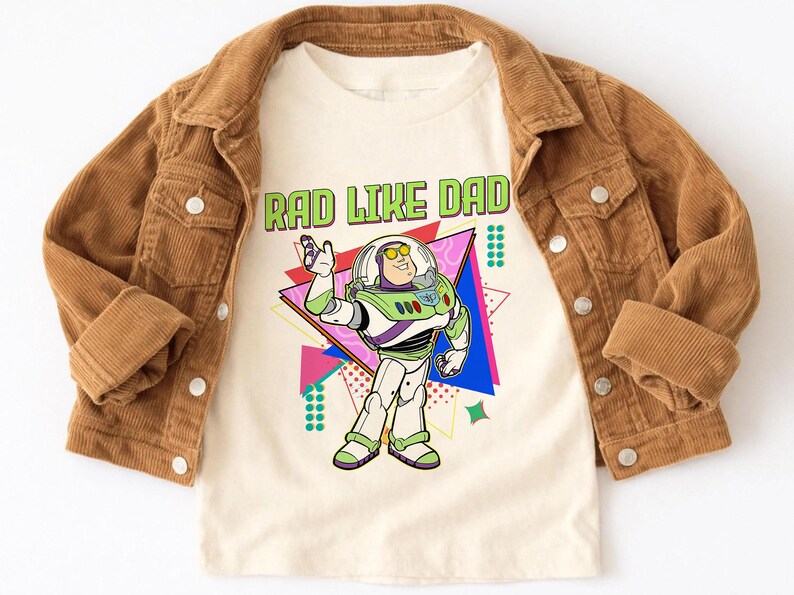 May include: A tan t-shirt with the text "RAD LIKE DAD" in green and a cartoon image of Buzz Lightyear. The shirt is layered under a brown corduroy jacket.
