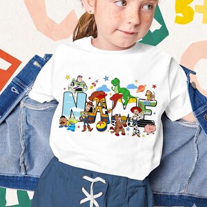 Puede incluir: Camiseta blanca con la palabra "NAME" en letras de bloque coloridas, cada letra adornada con personajes de dibujos animados. El dise&ntilde;o incluye personajes como Buzz Lightyear y Woody. Usada por una ni&ntilde;a.
