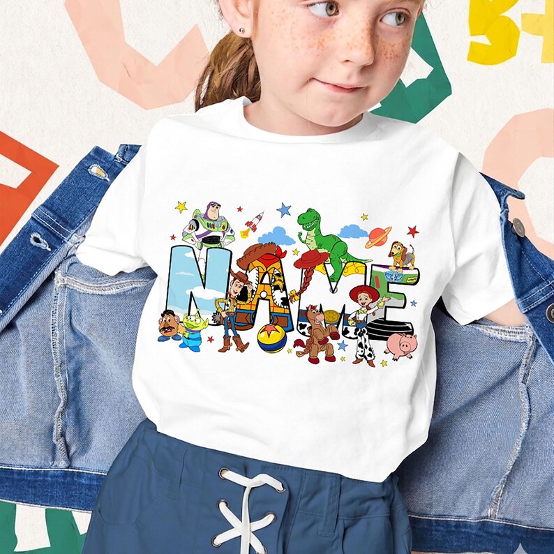 Puede incluir: Camiseta blanca con la palabra "NAME" en letras de bloque coloridas, cada letra adornada con personajes de dibujos animados. El dise&ntilde;o incluye personajes como Buzz Lightyear y Woody. Usada por una ni&ntilde;a.