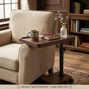 May include: A dark brown, adjustable side table with a rectangular top, resting over a beige armchair. A mug, book, and glasses sit on the table. The table has a black, adjustable pole and a square base with wheels. The text at the bottom says "DECENT CRAFTSMANSHIP - YOUR COZY LIFE".