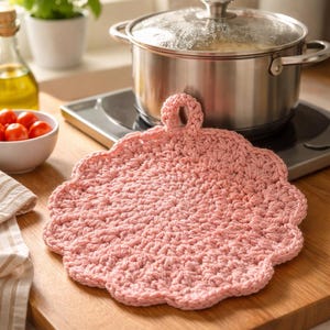 May include: A pink, crocheted potholder with a scalloped edge and a loop for hanging. The potholder is placed on a wooden cutting board next to a stainless steel pot on a stovetop. The kitchen setting includes a bowl of tomatoes and a bottle of olive oil.