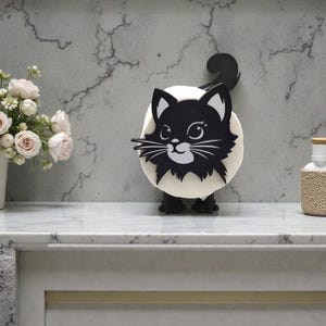 Puede incluir: Un objeto decorativo con forma de gato en blanco y negro. El gato tiene una cara negra con bigotes y orejas blancas, y una cola negra. Está sobre una base blanca con patas negras. A la izquierda hay un jarrón con rosas rosas.