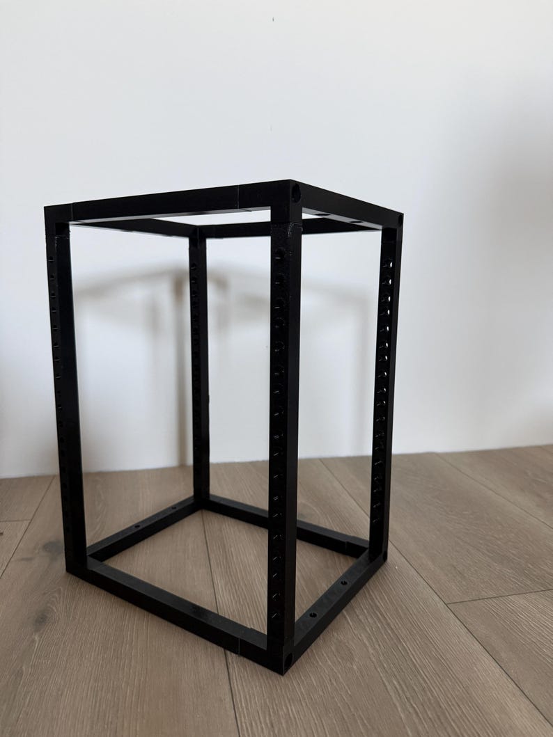 May include: A black rectangular frame constructed from four vertical pillars and eight horizontal connecting bars. The vertical pillars feature a series of evenly spaced circular holes along their length, creating a modular open-structure rack.