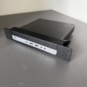 Może przedstawiać: Czarny minikomputer HP EliteDesk zamontowany w czarnym wsporniku do szafy serwerowej wydrukowanym w 3D. Przedni panel posiada srebrną płytkę z dwoma portami USB, portem USB-C, gniazdami audio i przyciskiem zasilania, w czarnej teksturowanej ramce.
