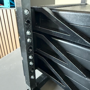 May include: A black 3D-printed rack frame featuring a vertical support beam with multiple hexagonal holes and silver metal screws. The structure includes angled side supports connecting to a solid black rectangular component within the frame.