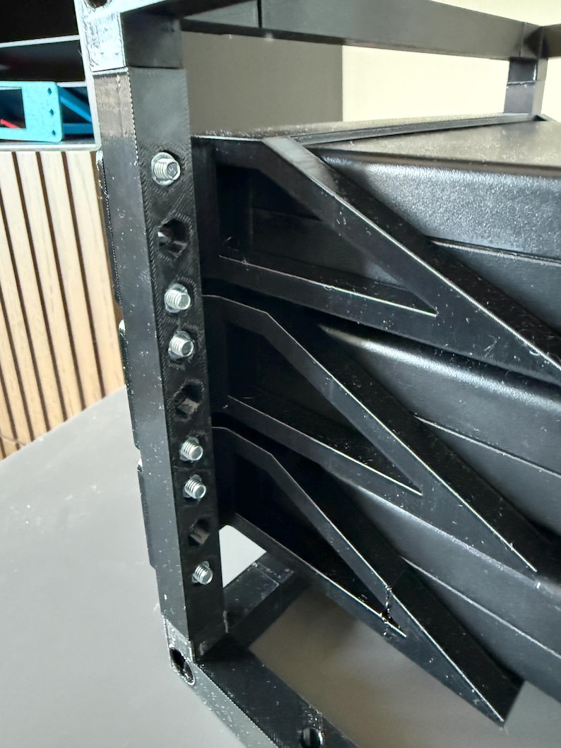 May include: A black 3D-printed rack frame featuring a vertical support beam with multiple hexagonal holes and silver metal screws. The structure includes angled side supports connecting to a solid black rectangular component within the frame.