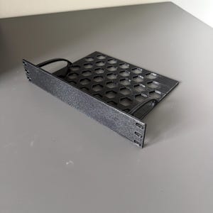 Op de afbeelding: Een zwarte rack-mount plank met een plat voorpaneel met drie montagegaten aan elke kant en een horizontale bodemplaat met een herhalend zeshoekig uitgesneden patroon voor ventilatie, gemaakt van een getextureerd materiaal.