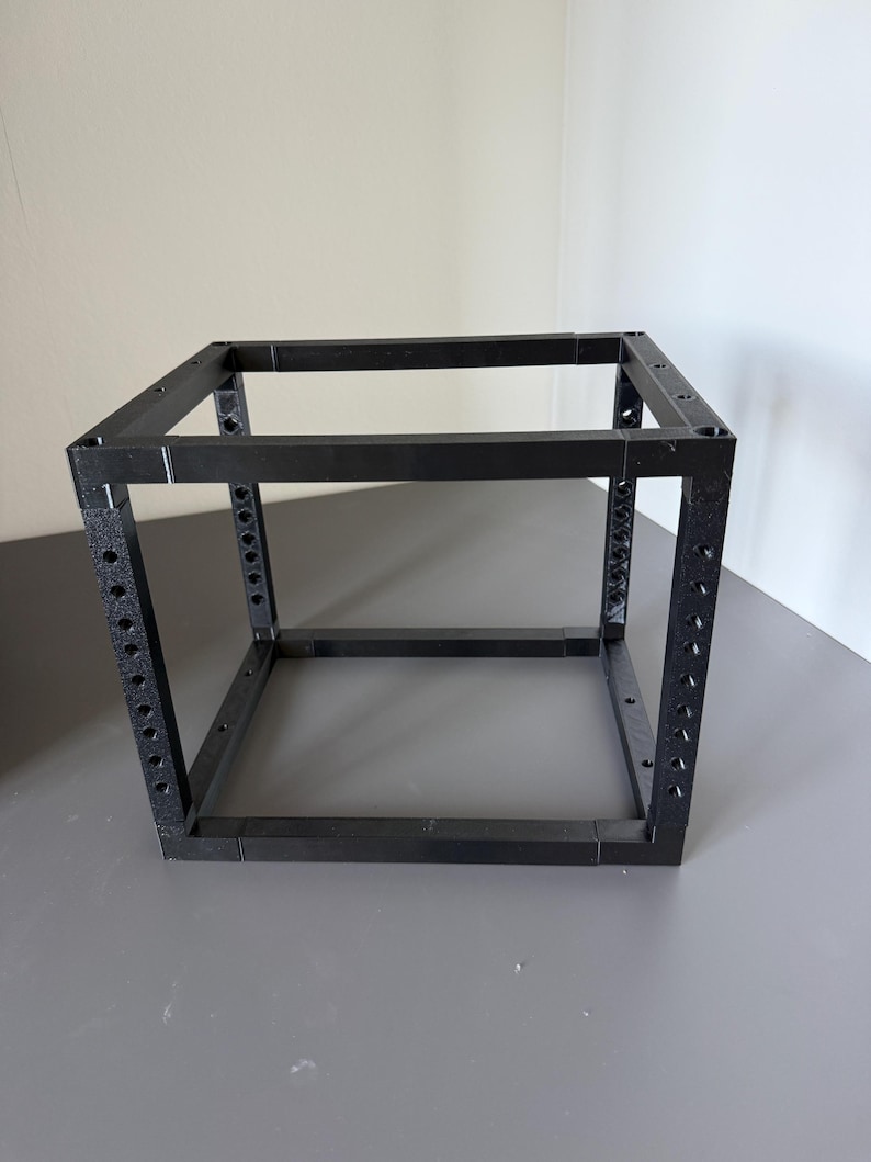 May include: A black, open-frame cube structure constructed from modular components. The four vertical corner posts feature a series of evenly spaced circular holes along their length, while the horizontal connecting bars are solid and smooth.