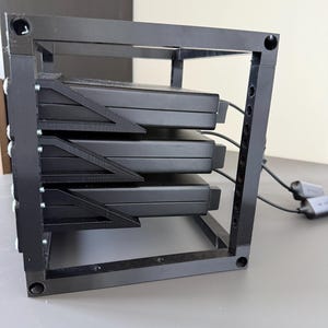 May include: A black, cubic, open-frame rack structure holding three stacked, rectangular black electronic devices. The frame features vertical supports with circular holes and triangular bracing, with black cables extending from the rear of the units.