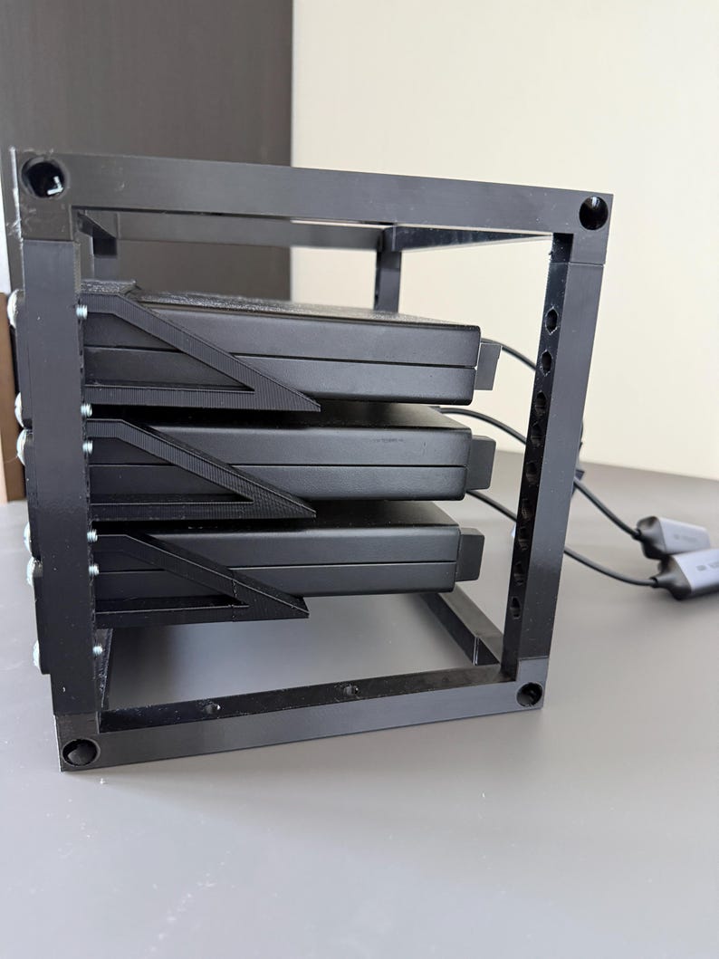 May include: A black, cubic, open-frame rack structure holding three stacked, rectangular black electronic devices. The frame features vertical supports with circular holes and triangular bracing, with black cables extending from the rear of the units.