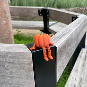 Op de afbeelding: Een klein, oranje, gearticuleerd pompoenfiguurtje met een groene steel. De pompoen heeft een rond, geribbeld lichaam, twee kleine armen die naar de zijkanten uitsteken en twee bungelende benen met voeten, allemaal met zichtbare gewrichten.