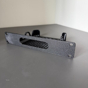 May include: A black, rectangular rack-mount bracket featuring a textured finish, a central horizontal cutout with a grid-like mesh pattern, and two circular mounting holes on each side, along with two vertical support tabs extending from the rear.