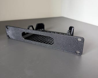 May include: A black, rectangular rack-mount bracket featuring a textured finish, a central horizontal cutout with a grid-like mesh pattern, and two circular mounting holes on each side, along with two vertical support tabs extending from the rear.