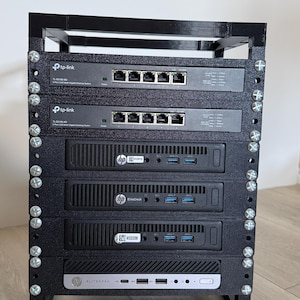 Op de afbeelding: Een zwart 3D-geprint rek met twee TP-Link TL-SG105-M2 5-poorts 2.5G multi-gigabit switches en vier HP EliteDesk mini-pc's. De componenten zijn verticaal gestapeld en vastgezet met zilverkleurige metalen schroeven in het frame.