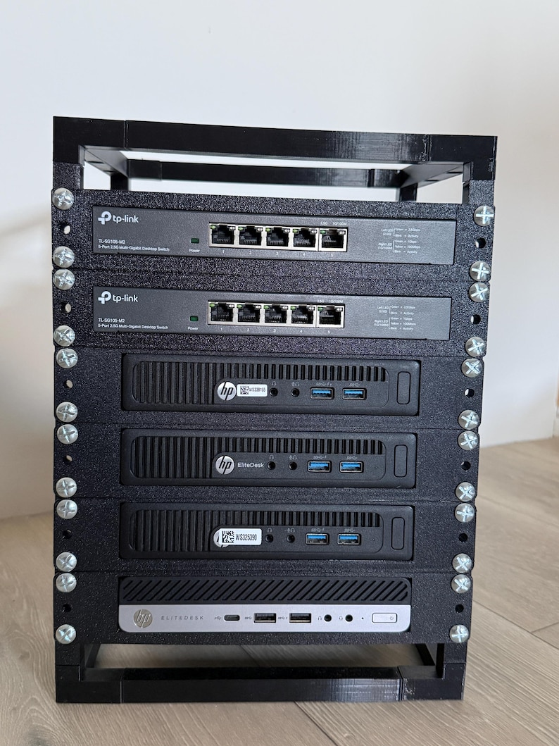May include: A black 3D-printed rack housing two TP-Link TL-SG105-M2 5-port 2.5G multi-gigabit desktop switches and four HP EliteDesk mini PCs. The components are stacked vertically and secured with silver metal screws in the frame.