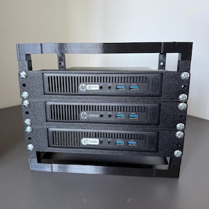 May include: A black 3D-printed rack mount frame holding three HP EliteDesk mini computers. The frame features vertical side supports secured with silver metal screws. Each computer unit displays the HP logo, front-facing USB ports, and audio jacks.
