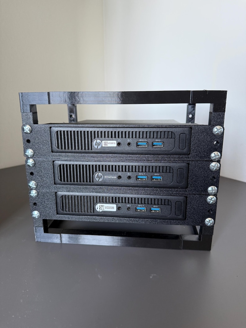 May include: A black 3D-printed rack mount frame holding three HP EliteDesk mini computers. The frame features vertical side supports secured with silver metal screws. Each computer unit displays the HP logo, front-facing USB ports, and audio jacks.
