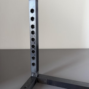 May include: A black 3D-printed frame structure featuring a vertical post with a series of evenly spaced hexagonal holes. The frame is constructed from interconnected square-profile bars joined at a right-angle corner, showing a textured finish.