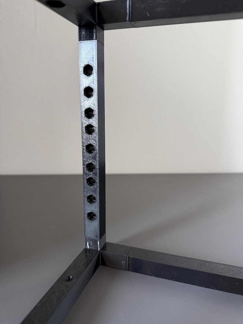 May include: A black 3D-printed frame structure featuring a vertical post with a series of evenly spaced hexagonal holes. The frame is constructed from interconnected square-profile bars joined at a right-angle corner, showing a textured finish.