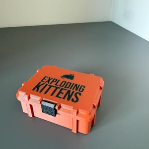 Könnte beinhalten: Ein rechteckiger Aufbewahrungskoffer aus orangefarbenem Kunststoff mit einem schwarzen Verschluss und dem auf den Deckel gedruckten Text "EXPLODING KITTENS", begleitet von einem kleinen schwarzen Katzensymbol über dem Schriftzug. Das Gehäuse ist texturiert.
