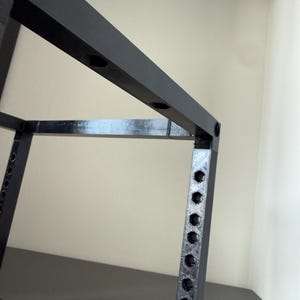 May include: A black, 3D-printed frame structure featuring a series of evenly spaced hexagonal holes along its vertical and horizontal beams. The object has a matte, textured finish and a rigid, geometric design with sharp, right-angled corners.