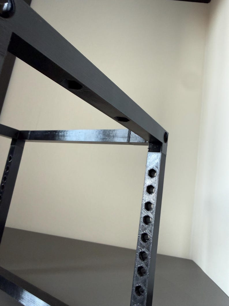 May include: A black, 3D-printed frame structure featuring a series of evenly spaced hexagonal holes along its vertical and horizontal beams. The object has a matte, textured finish and a rigid, geometric design with sharp, right-angled corners.