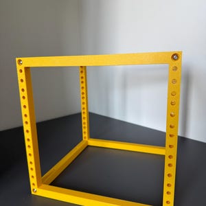 May include: A bright yellow, open-frame cube structure constructed from four vertical pillars and four horizontal bars. Each vertical pillar features a series of evenly spaced circular holes along its length, with small metallic fasteners at the corners.