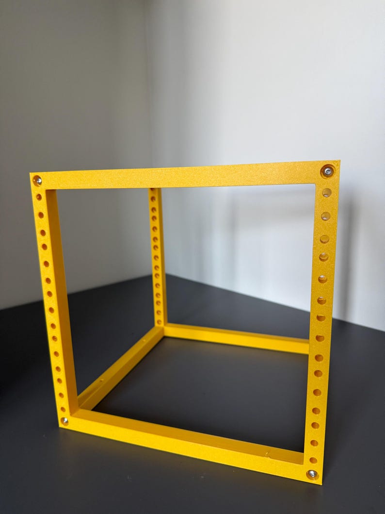 May include: A bright yellow, open-frame cube structure constructed from four vertical pillars and four horizontal bars. Each vertical pillar features a series of evenly spaced circular holes along its length, with small metallic fasteners at the corners.