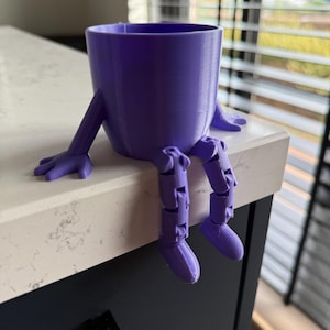 Op de afbeelding: Een paarse 3D-geprinte plantenbak in de vorm van een klein figuurtje met een cilindrisch lichaam, twee beweegbare armen die op het oppervlak rusten en twee bungelende benen met scharnierende knieën en voeten over de rand.