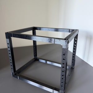 May include: A black, 3D-printed cubic frame structure featuring four vertical corner posts with evenly spaced circular holes along their length, connected by horizontal bars at the top and bottom to form an open, hollow box shape.