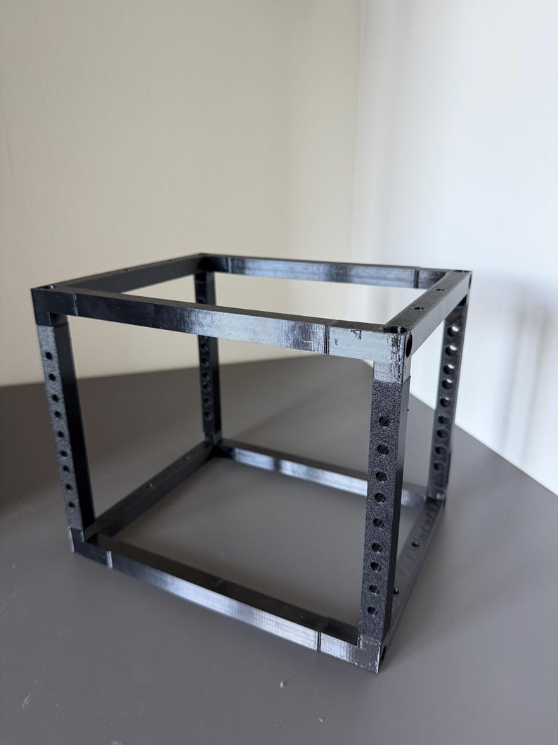 May include: A black, 3D-printed cubic frame structure featuring four vertical corner posts with evenly spaced circular holes along their length, connected by horizontal bars at the top and bottom to form an open, hollow box shape.