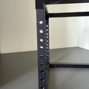 May include: A black metal rack frame featuring a vertical support beam with a series of evenly spaced circular holes along its length. The structure has a textured, matte finish and consists of connected square-profile metal bars.
