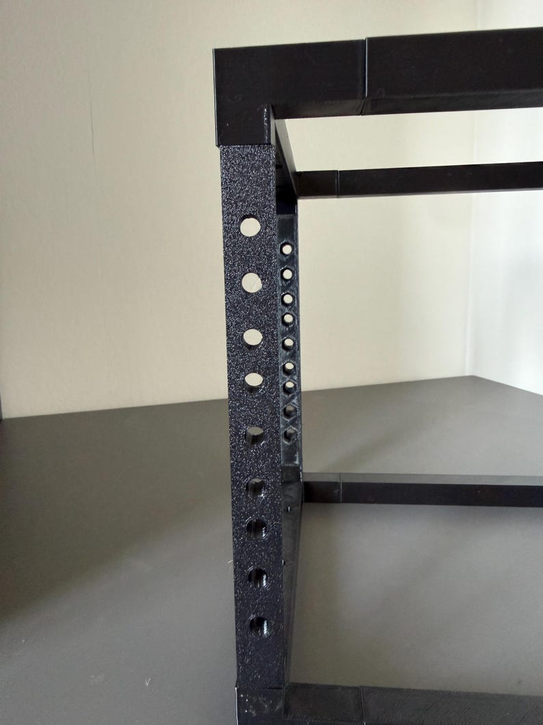 May include: A black metal rack frame featuring a vertical support beam with a series of evenly spaced circular holes along its length. The structure has a textured, matte finish and consists of connected square-profile metal bars.