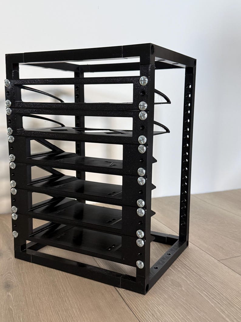 May include: A black modular storage rack featuring a vertical frame with five horizontal shelves. The structure is assembled with silver-colored metal screws visible along the side panels, creating a grid-like appearance for organizing items.