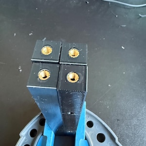 May include: Four black rectangular plastic components arranged in a square formation, each featuring a centred brass threaded insert at the top. The components are held upright by a blue plastic support structure with circular cutouts.