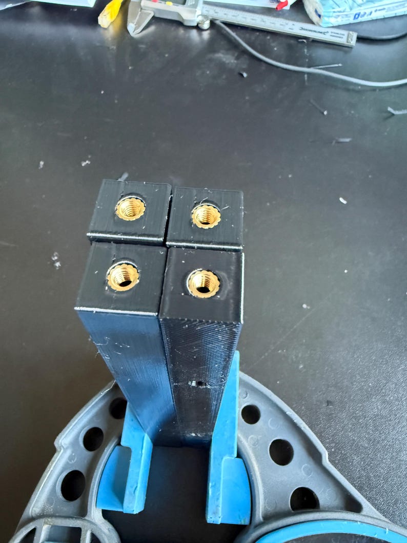 May include: Four black rectangular plastic components arranged in a square formation, each featuring a centred brass threaded insert at the top. The components are held upright by a blue plastic support structure with circular cutouts.