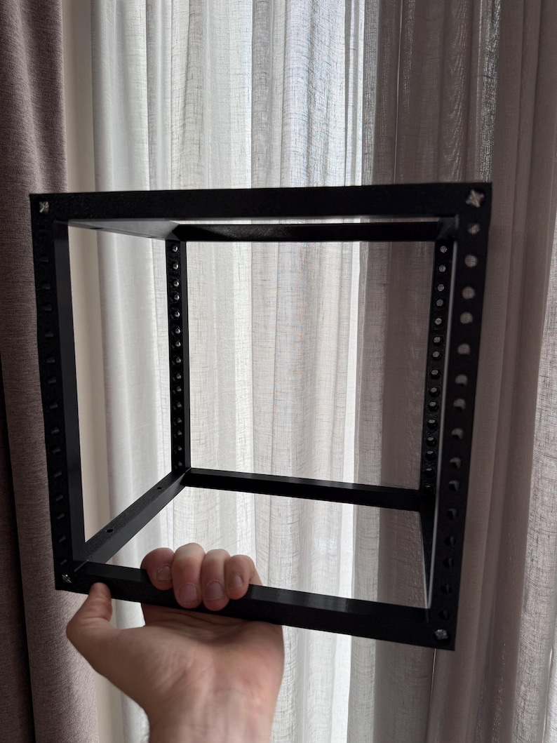 May include: A black, open-frame cube structure constructed from four vertical rails and four horizontal bars. Each vertical rail features a series of evenly spaced circular mounting holes along its length, connected at the corners by metal fasteners.