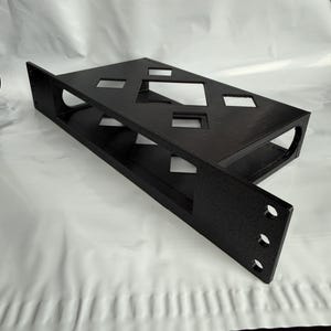 Pode incluir: Uma prateleira de montagem em rack preta com acabamento texturizado, uma base retangular plana com vários recortes em forma de diamante e um painel frontal vertical com três orifícios de montagem circulares e um grande recorte retangular.