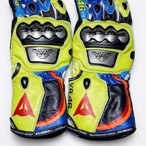 Puede incluir: Par de guantes de moto amarillos y negros con detalles azules y blancos. Presentan un gran logotipo "A" rojo, el número "46" y el texto "VR 46". Incluye protectores de nudillos plateados y logotipos de marca.