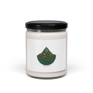 Op de afbeelding: Een witte kaarsenpot met een zwart deksel. Het etiket toont een groene, oranje en blauwe bergafbeelding met een kompasroos en coördinaten. Een woondecoratie-artikel.
