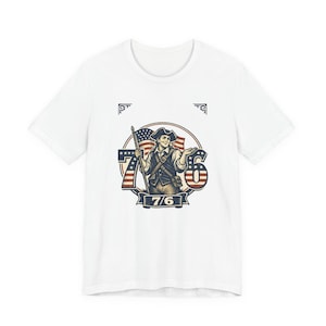 Op de afbeelding: Wit T-shirt met een patriottisch ontwerp. De afbeelding toont een koloniale man met een vlag en de cijfers "76" in rood, wit en blauw. De tekst "7/6" staat onderaan het ontwerp. Het shirt heeft een klassieke ronde hals.
