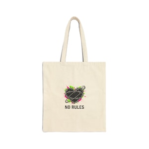 Puede incluir: Bolso tote color crema con un corazón negro atravesado por un imperdible, rodeado de salpicaduras de pintura verde y rosa. Las palabras "NO RULES" están impresas debajo. El bolso tiene asas largas.