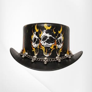 May include: A black top hat featuring a graphic print of three white skulls surrounded by yellow and orange flames. A silver-toned metal chain is wrapped around the base of the crown, adorned with small skull charms spaced along the chain.
